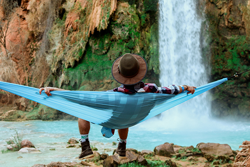 A person relaxing on a blue hammock with a waterfall in the background.